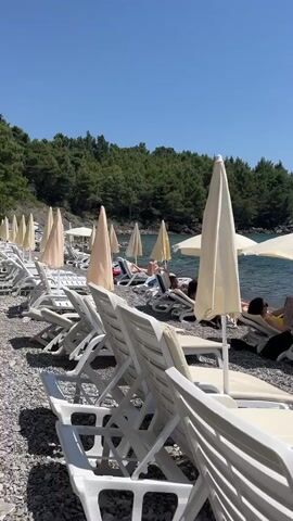 This video showcases a serene beach scene in Zagrađe, Montenegro. The primary subjects are people relaxing on lounge chairs under the warm sun. Several individuals are visible, some lying down, others seated, enjoying their time by the sea. The objects in focus include numerous white plastic lounge chairs, some equipped with cream-colored cushions. Beach umbrellas, predominantly cream-colored, provide shade, with some open and others closed. The beach itself is composed of pebbles, leading to the clear, blue water of the sea.

The activity taking place is leisurely relaxation at the beach. People are sunbathing, unwinding, and enjoying the coastal environment. In the background, several boats are visible on the water, including a sailboat with its mast up and smaller motorboats. The sea appears calm and inviting, reflecting the clear blue sky. Lush green trees cover the hills that form the backdrop to the beach, suggesting a natural and somewhat secluded setting.

Notable details include the bright, sunny weather, indicating it's likely a warm day, possibly midday given the strong sunlight. The overall atmosphere is one of tranquility and vacation enjoyment. The clear sky and calm water contribute to a peaceful ambiance. There is no visible text in the video.