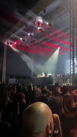 The video shows a crowd of people at an outdoor concert. A DJ is performing on stage, illuminated by bright lights and smoke machines. The audience is visible in the foreground, with some individuals dancing and cheering. The stage is set up with equipment, including turntables and speakers, and there are large screens displaying visuals. The lighting is dynamic, shifting between bright white and colored spotlights. The overall atmosphere appears energetic and lively, typical of a music festival or concert event.
