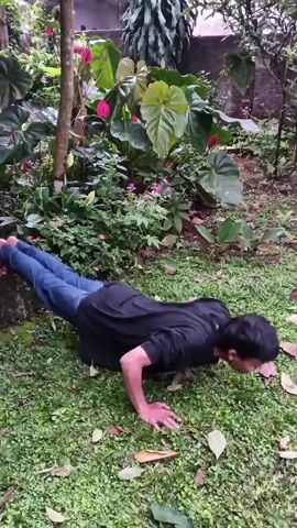 A man is attempting to do a push-up in a garden. He is wearing a black shirt and blue jeans. His feet are propped up against a small tree trunk. He lowers himself down, then loses his balance and falls forward onto the grass. The garden is lush with green ground cover, various plants, and pink flowers.