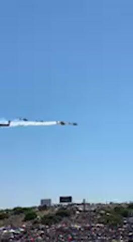 The video captures an outdoor scene under a clear blue sky, likely during a daytime event. A formation of at least five helicopters flies horizontally across the frame from left to right. One of the leading helicopters, on the far left, is emitting a distinct white smoke trail. Below the aerial display, a large crowd of spectators is visible in the foreground, partially out of focus, suggesting a public gathering such as an airshow or parade. A multi-speaker sound system mounted on a pole is also present in the lower right of the frame. The weather appears bright and sunny. The event is taking place in Jerez de la Frontera, Spain. No discernible text is clearly visible within the frame.