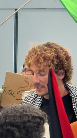 The video depicts a gathering of individuals, likely a protest, taking place in a public area, possibly an airport terminal. The primary focus is on a man with curly hair, wearing a black and white patterned jacket, who appears to be speaking or chanting. He is surrounded by other people holding signs and flags, including the Palestinian flag. A sign visible in the video has the words "Total S" on it. People are recording the event with their phones and cameras. There is a sign visible that reads "Votre taxi officiel vous attend Porte 24" (Your official taxi awaits you at Gate 24). Law enforcement personnel are also present in the background.