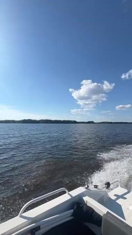 The video opens with a view of the water from a boat, with a bright sun shining in a clear blue sky. As the boat moves, the wake behind it is visible, along with the shoreline in the distance. Fishing rods are set up on the boat. The sun is bright, suggesting it is daytime. The water appears calm with some ripples. The scene is a scenic view of a body of water, possibly a lake or a wide river, with a treeline on the horizon. The boat itself is white with some metal railings and a windshield. A "LOWRANCE" branded electronic device is visible on the console of the boat. The location is identified as Syukterka, Russia. The overall mood is peaceful and relaxing, evoking a sense of enjoying a day out on the water.
