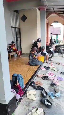The video shows an outdoor scene with a walkway or porch area. Several people are present. On the left side, a young boy is sitting on a bench, looking at a yellow object. A motorcycle is parked behind him. Further to the right, a woman wearing a hijab and a young boy are sitting on the ground. Many pairs of shoes, mostly sandals and flip-flops, are scattered on the ground in front of them. A few motorcycles are parked along the side of the building. In the background, there is a street with a few more people and motorcycles. Two young girls and a person wearing a helmet are standing near a motorcycle on the right side of the scene. The environment appears to be a residential or communal area, with buildings and a street visible. There are also flags visible, suggesting a possible event or celebration.