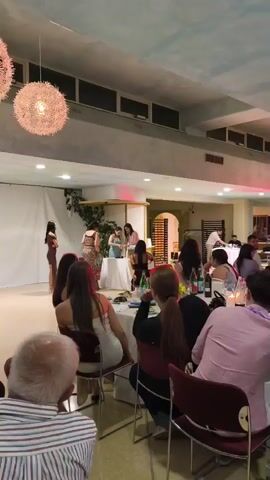 A video shows an indoor event with several people. The setting appears to be a banquet hall or restaurant, with tables set for dining. In the foreground, a group of people are seated at tables facing a performance area. In the background, on a raised platform, a group of women are gathered around a table. One woman on the left side of the platform is wearing a brown dress and appears to be dancing or performing. Others on the platform are dressed in what appear to be formal or semi-formal attire. The room is lit by overhead lights, and decorative light fixtures are visible hanging from the ceiling. The walls are painted a light color, and there is a white curtain or backdrop on the left side of the performance area. A doorway or archway is visible in the background on the right side of the frame. The overall atmosphere suggests a celebratory occasion, possibly a party or event.