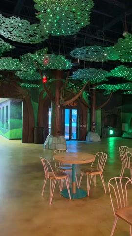 The video pans across an indoor dining area designed with a whimsical forest theme. The ceiling is adorned with large, stylized green tree canopies, each dotted with numerous small green lights, creating a starry, ethereal effect. Several large, artificial tree trunks are strategically placed throughout the space, some curving to support the ceiling canopy. The walls are painted with a mural depicting a lush, green landscape, enhancing the forest ambiance. In the foreground, light-colored, round tables with teal bases are surrounded by pink wire chairs with wooden seats, arranged in small clusters. In the background, a darker, arched entryway is visible, also decorated with green lights. To the right of this entryway, a series of small, stall-like structures with peaked roofs are lined up, suggesting a food court or service area. Several digital screens are mounted on the walls, some displaying "ORDER HERE" signs. The floor is a polished, warm-toned material, possibly concrete or epoxy, reflecting the ambient light. The overall impression is that of a child-friendly, themed restaurant or entertainment venue. There are no people clearly visible in the frame.