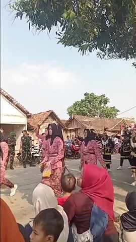 This is a vertical video of a procession or parade on a street in a village. In the foreground, several people are watching the event. The majority of the crowd is obscured by the bottom edge of the frame, but a few faces are visible, including those of children and adults wearing hijabs. The procession itself is in the mid-ground and background. On the left side of the street, there are several people dressed in green military-style uniforms. Further into the procession, a group of people are dancing or performing. Several of these individuals are wearing black outfits adorned with gold and black trim, and some have elaborate headdresses. There are also women in the procession wearing pink, patterned clothing and hijabs. In the background, there are simple village houses with tiled roofs. A few motorcycles are parked along the side of the street. Trees with green leaves and what appear to be mangoes are visible in the upper part of the frame. The sky is bright and appears to be daytime. The overall atmosphere suggests a cultural celebration or event.