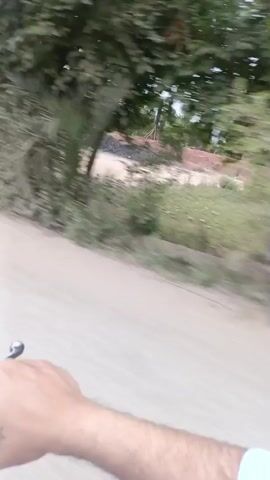 The video shows a view from a moving vehicle traveling on a dirt road. The arm of the person filming is visible in the lower left corner of the frame. The scenery consists of trees and green vegetation along the side of the road. In the distance, there is construction equipment, including what appears to be a tractor and some scaffolding structures. There are piles of dark material, possibly dirt or gravel. The sky is overcast, and the overall environment appears rural.