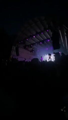 A live concert is in progress. A crowd of people is gathered in front of a stage. The stage is illuminated with purple lights. On the stage, a musician is performing. Behind the musician is a circular backdrop with concentric rings. The overall atmosphere is dark, with the stage lights providing the primary illumination.