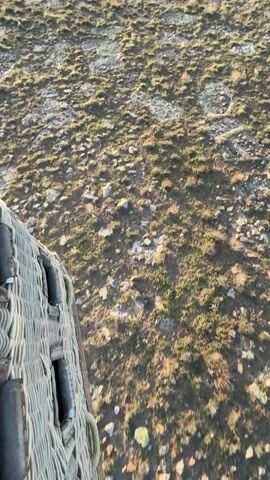 The video opens with a close-up view from inside a hot air balloon basket, looking down at a rugged, dry landscape covered in sparse vegetation and rocks. The sound of the hot air balloon's burner is audible. As the camera pans upwards, a breathtaking panoramic view unfolds. Several colorful hot air balloons are seen floating in the distance, dotting a clear blue sky. Below them, a large, calm body of water (likely a lake or reservoir) is nestled among rolling hills and mountains. The landscape is arid, with earthy tones of brown, green, and grey. The warm, soft light suggests either an early morning sunrise or a late afternoon sunset, casting long shadows and illuminating the scene beautifully. One hot air balloon in the mid-ground has text on its side, partially visible as "Tulip" or similar. The setting is a vast, open natural environment, characteristic of regions suitable for hot air ballooning, such as Yeşilhisar, Turkey.