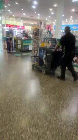 A person in a black shirt and black pants with a green armband pushes a cleaning cart through a brightly lit retail store. The cart contains cleaning supplies such as bottles and rags, as well as a mop. The person is seen from the side and slightly from behind, with their left hand on the cart's handle. The store appears to be a pharmacy or drugstore, with shelves of products visible in the background, including a display labeled "Revlon." The floor is a speckled terrazzo. The video is shaky, suggesting it was filmed handheld.