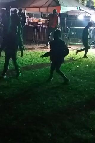 The video shows two individuals dancing on a grassy area, illuminated by color-changing spotlights. They are in silhouette, but their movements suggest they are facing each other. Behind them, a tent structure is visible, with several people standing underneath and around it. A metal barrier partially obscures the view. Another individual in a hat walks past the camera. The light keeps changing color, bathing the scene in alternating shades of green, blue, pink, and red. The setting appears to be an outdoor event or party.