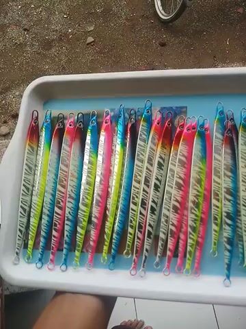 Here is a concise description of the video:

The video shows a white plastic tray filled with around twenty brightly colored fishing lures partially submerged in water. The lures are long, thin, and metallic, painted in a variety of vibrant hues including pink, blue, green, red, and silver, with a tiger-stripe pattern over most of them. The tray is being held up by a person whose feet are visible in the corner of the frame, wearing open-toed sandals.  The background is somewhat indistinct, showing part of a bicycle wheel and a tiled surface.