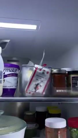The camera zooms into a refrigerator shelf. On the shelf, among other items, is a container holding a package of Fairview South African Feta with Garlic & Chilli. The refrigerator is lit by an overhead light.