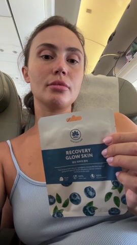 In an interior medium shot, a woman sits in an airplane seat, looking directly at the camera and holding up a sheet mask. She is fair-skinned, with dark hair pulled back. She is wearing a light blue tank top. The sheet mask packet is mostly white with blue trim, and features an image of blueberries. The text on the packet reads "RECOVERY GLOW SKIN." The airplane cabin is visible in the background. A small child's face peeks over the top of the seat behind her. The woman holds the sheet mask up with both hands. She appears to be demonstrating the product.