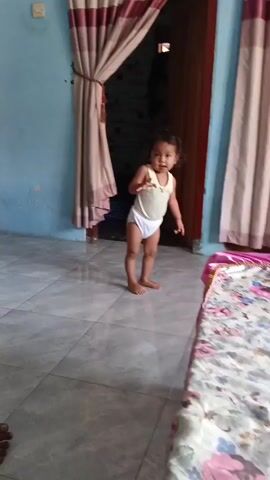 A young child, wearing a white diaper and a light yellow tank top, stands on a tiled floor. The child has dark, curly hair and is facing the camera. The child's left arm is extended towards the camera, with the hand open. Behind the child, there is a doorway with dark wood and a tan curtain hanging to the left. To the right of the child is a bed with a pink and floral patterned comforter. The walls appear to be painted blue and are somewhat aged. The lighting is dim, suggesting an indoor setting.
