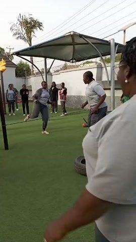 The video shows a group of adults and children playing in a playground. The ground is covered in green artificial turf. There is a metal structure with a canopy roof. Some people are running and jumping over a rope. One person is on a seesaw. There are several tires lying on the ground. A tall palm tree is visible in the background.