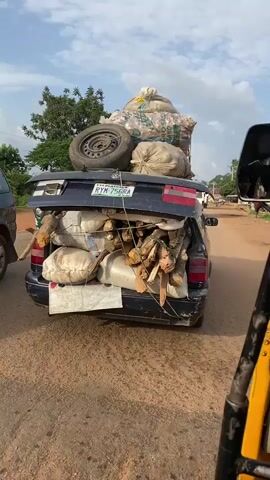 The media file shows a dark blue sedan overloaded with various items, being driven on a dirt road in Barikin Sabon Gidan Kanat, Nigeria. The car's trunk is open, and its contents are piled high, extending above the roof. Visible items include several large, white sacks, bundles of firewood, and a spare tire. One sack on top appears to be filled with a lighter-colored material, possibly grains or feed. The car's license plate is partially visible and reads "RYM 756RA."

To the left of the sedan, a green and yellow auto-rickshaw (also known as a "Keke Napep" in Nigeria) is visible, carrying at least two passengers. The perspective suggests the media was captured from another auto-rickshaw, seen in the foreground with yellow and black framing.

The scene takes place outdoors during daylight hours, under a partly cloudy sky. The ground is dusty, indicating a dry season or a road that is not paved. Trees and foliage are present in the background, suggesting a rural or semi-rural setting. The overall impression is one of heavy transport, likely for trade or personal use, highlighting resourcefulness in utilizing available transportation to its maximum capacity. No strong emotions are conveyed, but the image depicts a common sight of overloaded vehicles in many parts of Nigeria.