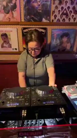 A woman with short, dark hair wearing glasses and a light green collared shirt stands in front of two Pioneer DJ consoles in a dimly lit room. She is operating the DJ equipment, with her hands on the decks, seemingly mixing music. Behind her, a wall is decorated with framed vinyl record covers featuring artists like Freddie Mercury, Bob Marley, and others. A red "ON AIR" sign is visible at the bottom of the frame. There are CDs stacked to the right of the DJ equipment. People can be heard cheering in the background.