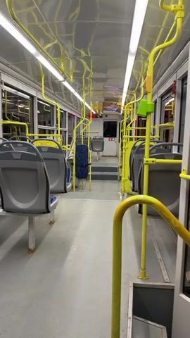 The video shows the interior of a public bus or tram. The camera is positioned in the aisle, looking towards the back. The seats are gray and appear to be empty. Yellow handrails and stanchions are visible throughout the cabin, as are fluorescent lights on the ceiling. There is a set of stairs leading to another section of the vehicle, and a door is visible at the end. The overall impression is that of a clean, well-lit, and empty passenger vehicle.