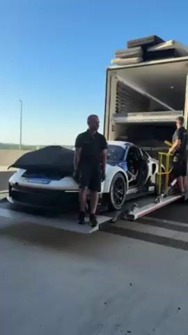 Here is a description of the video:

The video shows a white and blue race car being unloaded from a large white truck. The car is sitting on a hydraulic lift, and two men are present. One man, wearing a black shirt and shorts, is standing near the car, leaning on the lift and looking toward the camera with a smile. The other man, who is partially obscured, is operating the lift with a yellow control panel. The truck is parked on a paved surface, with a view of greenery and the sky in the background. There is music playing in the background.