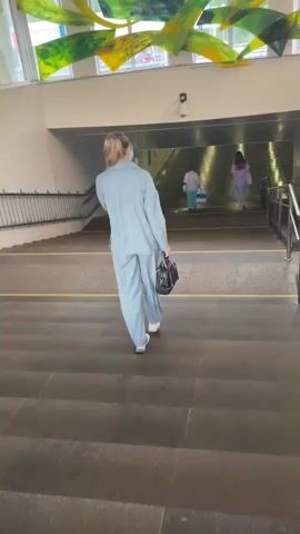 A woman in a light blue pantsuit walks up a concrete staircase towards a brightly lit underpass. She carries a black handbag and wears white sneakers. Her blonde hair is pulled back in a bun. In the underpass, two figures are visible, one in a white top and blue shorts, the other in a purple top and light-colored skirt. They are also moving away from the viewer, towards the other side of the underpass. The underpass appears to be a pedestrian walkway, with railings on either side of the stairs. Above the underpass entrance, a colorful abstract art installation made of wavy green and yellow panels is suspended from the ceiling. The overall scene suggests daytime, and the lighting in the underpass indicates it is well-lit. The setting appears to be an urban environment, possibly a subway station or a pedestrian underpass connecting different parts of a city. There is no visible text in the image.