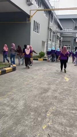 A group of women, most wearing headscarves and casual attire, are walking away from the camera on a paved outdoor area. The building behind them appears to be a commercial or industrial facility, with a gray facade, windows, and some external piping and structures. Some women are walking in a line, while others are more spread out. The ground is concrete with painted yellow lines.