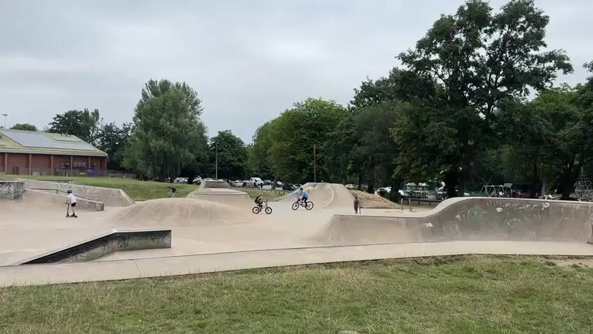 A wide shot of a skatepark on a cloudy day. The skatepark is made of concrete and features various ramps, bowls, and ledges. Several people are present, including a few on skateboards and others on BMX bikes. In the background, there is a building with solar panels on the roof, trees, and a parking lot with cars. The ground in the foreground is covered with grass. The camera is static and pans slowly from left to right.