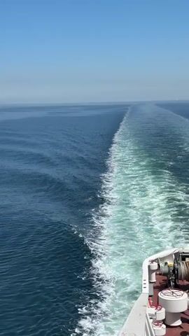 The video shows a view from the back of a ship, looking out over a calm, blue sea under a clear, light blue sky. The ship leaves a white foamy wake behind it. In the distance, the horizon is visible with a very small object on the water. A larger object, presumably another ship, is further away.