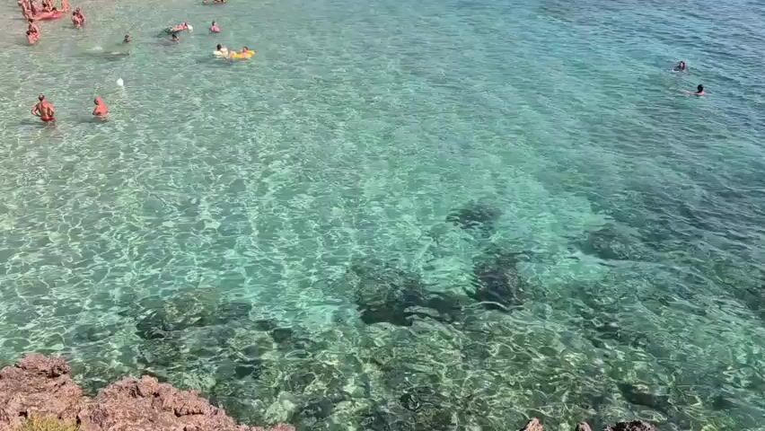 The video shows a view of a clear turquoise ocean with people swimming and floating. The water is so clear that the rocky bottom is visible. On the left side of the frame, several people are in the water, some swimming and one person on an inflatable float. Further out, two individuals are swimming. In the upper left corner, a white buoy is visible. The foreground features a rocky shoreline with some sparse vegetation. The scene depicts a sunny day at the beach.