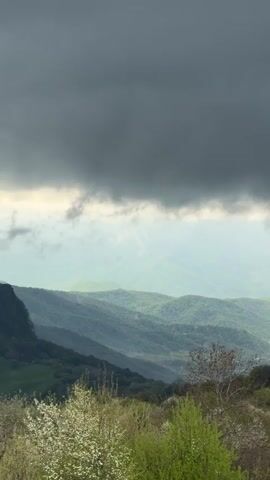 The video shows a mountainous landscape under a heavy, dark gray cloud cover. The foreground has green trees and shrubs, some with white blossoms. The mid-ground features rolling green hills disappearing into a hazy distance. The sky is dominated by the ominous cloud formation, with a sliver of light peeking through. There are no people or animals visible in the scene. The video appears to be taken from a stationary position, possibly from an elevated location overlooking the valley.