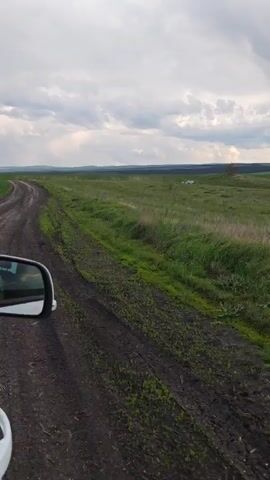 The video shows a view from inside a white car driving on a dirt road. The road runs alongside a large, grassy field. The sky is overcast with clouds, suggesting a potentially rainy or stormy day. The car's side mirror is visible, reflecting the surrounding landscape. The overall scene is rural and gives the impression of traveling through open countryside.