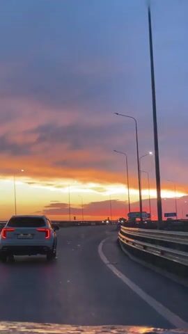A rear view shot from inside a vehicle shows the road ahead during a sunset. The sky is a gradient of deep blue, pink, and orange. Several streetlights line the right side of the road. A white SUV is in front, in the right lane, its brake lights on. Another white car is in the left lane, further ahead. Traffic signs are visible, including one green sign with white text and arrows. The road appears wet, reflecting the headlights of the vehicles.