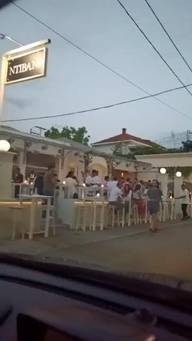 The video shows a street scene at dusk, possibly in a Mediterranean town. Several outdoor seating areas of restaurants or cafes line the street, with people seated at tables and standing. There are cars driving on the street, and some people are walking. The overall atmosphere appears to be lively and social, suggesting a warm evening. The architecture of the buildings appears to be white with some terracotta roofs. The lighting suggests it is twilight or early evening, with some artificial lights illuminating the seating areas and street.