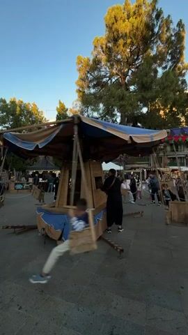Here’s a description of the video:
The video shows a carousel in motion. It is built of wood and covered by a blue and brown canvas. The base of the carousel is also blue and brown, resembling a boat. Children are riding in wooden boxes suspended by ropes from the top of the carousel. A person in black clothes is turning a crank that rotates the carousel. People are milling around in the background, with tents and tables set up, suggesting a fair or market atmosphere. A large tree can be seen in the background, along with a clear, blue sky.