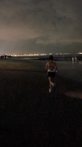 The video shows a person running on a dark sandy beach towards the ocean at night. In the background, there are scattered lights of a city visible across the water. Other people are distantly seen on the beach. The sky is overcast, contributing to the dimness of the scene.