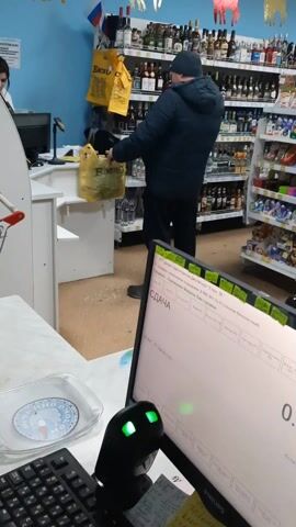 The video shows a man paying for beer at a checkout counter in a store. The man, wearing a dark jacket and hat, stands at the counter holding a yellow shopping bag filled with beer bottles. He is paying the cashier with cash. The cashier stands behind the counter, visible from the shoulders up. Behind the man are shelves stocked with various bottles, likely alcohol. The wall behind the shelves and checkout area is blue with horse silhouettes. A computer monitor displays transaction information. A shopping cart is visible to the left of the counter.