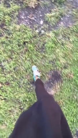 The video begins with a first-person perspective of someone walking on a grassy, uneven surface. The camera then pans up to show a bright blue sky with some wispy clouds. The perspective shifts to a vertical orientation, showing a young woman with long brown hair looking up at the sky. She is wearing a black hoodie. In the background, there is a wooden fence, a grassy field, and distant mountains. Some cows are visible in the field behind the fence. The sun appears to be setting, casting a warm light on the scene.