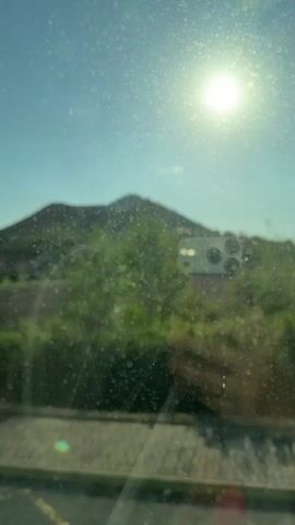 This is a first-person perspective video of a vehicle moving along a road with a scenic view of a hill and the sun. The video begins with a view of a hill with a water tower on top and a building on its slope, seen through a dirty window. The vehicle turns a corner, revealing more of the landscape. The sun is bright and high in the sky, creating lens flares on the window. The surroundings are predominantly green with trees and grassy areas. The video continues to show the moving landscape as the vehicle progresses along the road. The camera appears to be mounted inside the vehicle, looking out the window.
