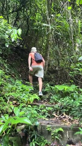 A person wearing a white hat, an orange t-shirt, and light-colored shorts with a black backpack is ascending a stone staircase in a lush, green forest. The stairs are partially covered in leaves and moss, and dense foliage lines both sides of the path. Sunlight filters through the canopy, illuminating the scene.
