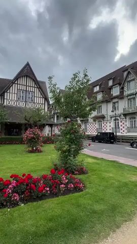 The video pans across a lush green lawn adorned with vibrant red and pink flowers in garden beds. Several trees, some with blooming pink flowers, are scattered throughout the lawn. In the background, a grand building with a distinctive Tudor-style facade, featuring dark timber beams against white walls, stands prominently. Adjacent to it, another section of the building displays a repeating pattern of red and white squares. A classic black vehicle is parked near the building. The sky overhead is overcast with dramatic clouds. The scene appears to be a well-maintained garden area in front of a historic hotel or large residence.