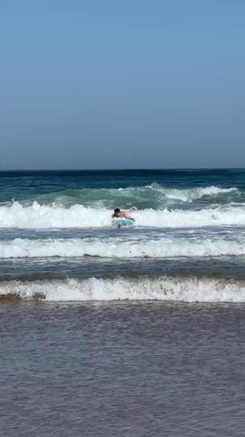 A person is seen on a boogie board in the ocean. The person is in the middle of a wave. The sky is clear and blue. The ocean is a deep blue with white waves. The person is wearing swim trunks. The person is holding onto the boogie board. The person is looking forward. The person is in the process of riding the wave.