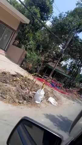 The video appears to be recorded from the perspective of a person riding a motorcycle. The footage is shaky and slightly blurred due to motion. In the foreground, a black side mirror of the motorcycle is visible. The scene depicts a street with buildings and trees. There are some red hoses laid out on the ground, possibly indicating some construction or utility work. In the background, a building with a green roof is visible, along with other structures and a few trees. The environment appears to be outdoors, possibly in a residential or commercial area. There are no clear individuals or specific actions identifiable due to the blurriness and the fast-paced nature of the recording. The video transitions between different views of the street as the motorcycle moves. In some frames, there are glimpses of more buildings and what might be market stalls or shops. There are also some white bags that could contain sand or other materials.