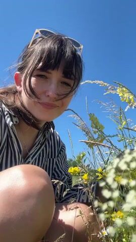 A woman with dark hair and bangs is kneeling in a field of wildflowers. She is wearing a striped shirt and sunglasses are on top of her head. The sky is blue and clear. The woman is looking at the flowers and appears to be smelling them. She is reaching out and touching some of the plants. The camera is positioned low to the ground, looking up at the woman.