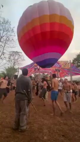 The video shows a group of people dancing around a large, colorful hot air balloon that’s tethered to the ground. The balloon has horizontal stripes of pink, orange, red, and purple. There is an open flame heating the air inside the balloon. The people are mostly bare-chested, and are dancing on a dirt ground. A man stands in the foreground holding what appear to be bamboo sticks. In the background, a colorful canopy structure is visible above the crowd.