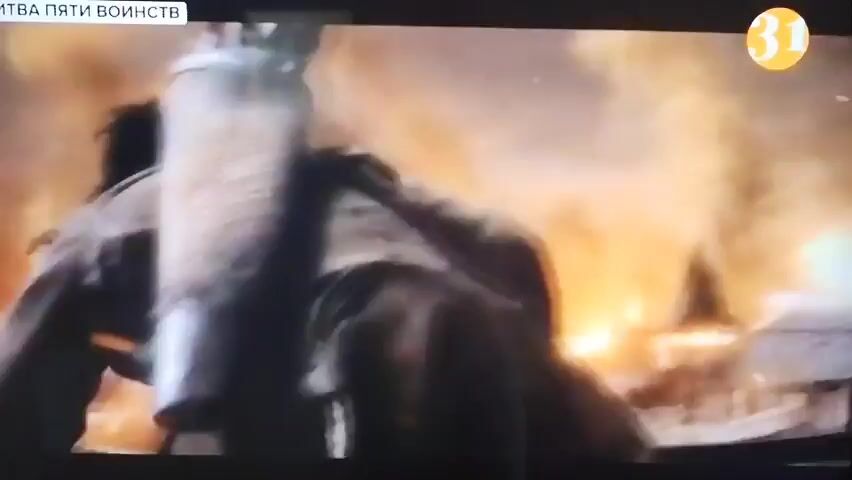 The video displays a cinematic scene of a village or town engulfed in flames. Buildings with wooden structures and tiled roofs are heavily ablaze, with large plumes of smoke and fire dominating the screen. The scene appears to be from a historical drama or fantasy film, possibly depicting a battle or raid. A small graphic overlay in the top left corner reads "БИТВА ПЯТИ ВОИНСТВ" which translates to "BATTLE OF THE FIVE ARMIES". Another graphic in the top right corner features a yellow circle with the number "31". There are no visible people in the frame. The overall ambiance is one of destruction and chaos.