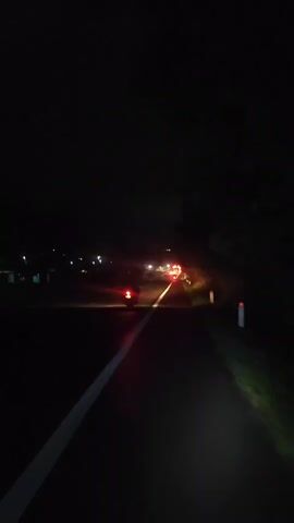 The video shows a dark asphalt road at night. There is a white line painted on the left side of the road. On the right side of the road, there is a white traffic post with a red band near the top. Trees and foliage are visible on the right side of the road. In the distance, there are lights from vehicles on the road, including red taillights and headlights. One vehicle appears to be a motorcycle with its taillight illuminated. There are also some faint lights in the background, possibly from buildings or streetlights. The overall scene is dimly lit and suggests a rural or suburban road at night.