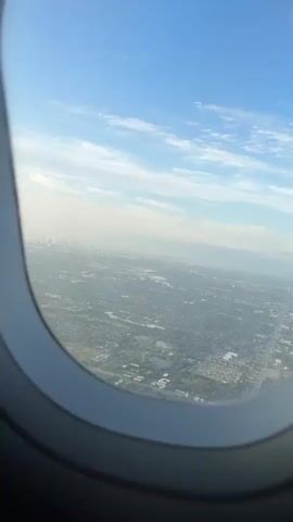 The video presents an aerial view of a sprawling urban and suburban landscape, seen through an airplane window. The camera looks out and slightly down, revealing a vast expanse of buildings, roads, and green spaces under a clear blue sky with scattered white clouds.

In the mid-ground and foreground, a dense network of residential and commercial structures is visible, interspersed with trees, parks, and what appear to be several lakes or large ponds. A prominent feature in the lower right portion of the frame is a large complex with vibrant blue pools, possibly a water park or resort. Straight lines of roads and highways cut through the developed areas.

On the distant horizon, a distinct city skyline rises faintly through a slight atmospheric haze, suggesting a major metropolitan area. The sun is illuminating the scene from the left, casting a soft light and contributing to the haze that somewhat obscures the furthest reaches of the cityscape. The overall impression is one of a vast, developed coastal region on a clear day, consistent with the provided location context of Dania Beach, United States of America, likely showing parts of South Florida. No discernible text is visible.
