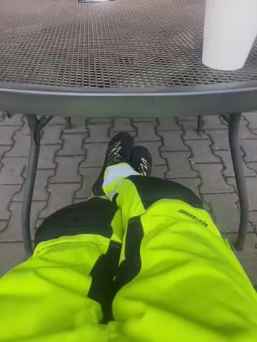 The video shows a first-person perspective of someone sitting at a round metal table with a mesh top. The person is wearing bright yellow pants with black accents and black athletic shoes. Their legs are crossed at the ankles. A white paper cup is on the table to the right. In the background, other people are visible sitting at the table, some holding cups. The ground is paved with interlocking bricks. The scene appears to be outdoors, possibly at a cafe or rest stop.