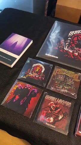 The video shows a table displaying merchandise from the band “The Pentagram Stringband.”  Merchandise includes vinyl records, CDs, cassettes, shirts, patches, ashtrays, lighter cases, and books. A handwritten price list is visible: "Records-7, Tapes-3, CDs-5, Shirts-20, Patches-3, Ashtrays-25, Lighter Cases-10." Ashtrays made of resin are displayed. The table is covered in black material.
