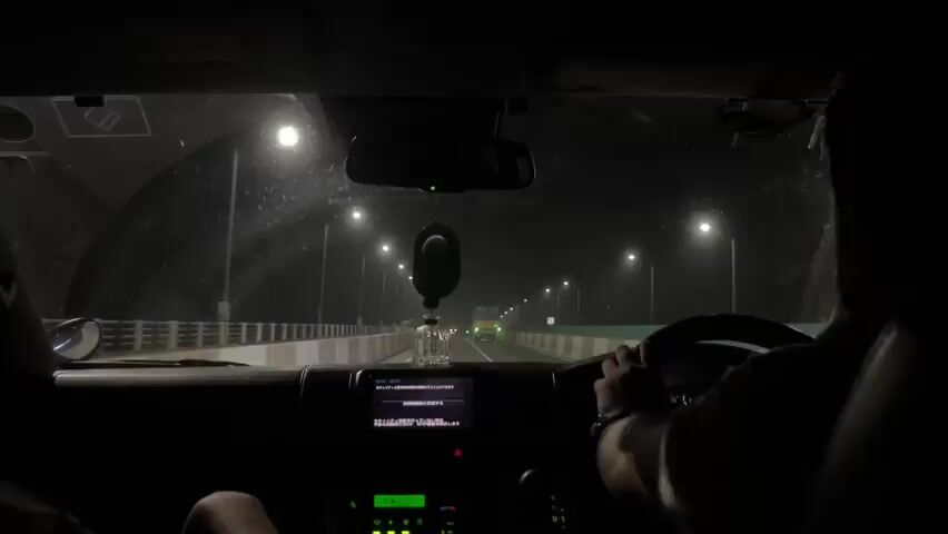 This is a POV shot from inside a vehicle at night, driving on a bridge over a body of water. The windshield is speckled with dirt and water. Streetlights line the bridge, casting a glow on the dark, rainy night. A dashboard with a navigation screen and radio is visible. A driver's hands are on the steering wheel. In the distance, other vehicles, including a large truck, are visible on the road. The interior of the vehicle is dimly lit.